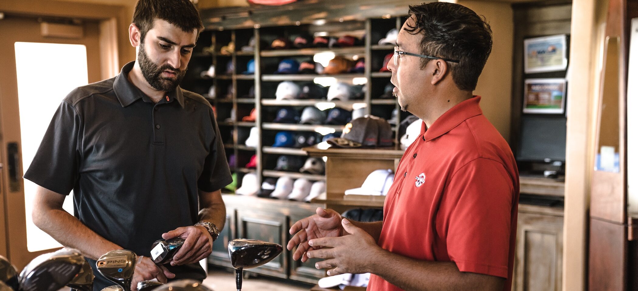 two people in a serious conversation about golf clubs and a shelf of hats in the background